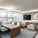 A modern looking office with hues of brown, white, and cream. Office includes a desk and chair, along with a sitting area space with a TV mounted on the wall.