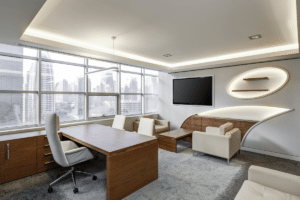 A modern looking office with hues of brown, white, and cream. Office includes a desk and chair, along with a sitting area space with a TV mounted on the wall.