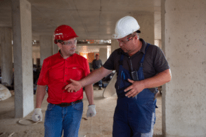 Two men in hard hats are talking while located on a construction site.