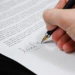 Image includes a person's hand holding a pen, signing a signature.