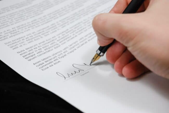 Image includes a person's hand holding a pen, signing a signature.